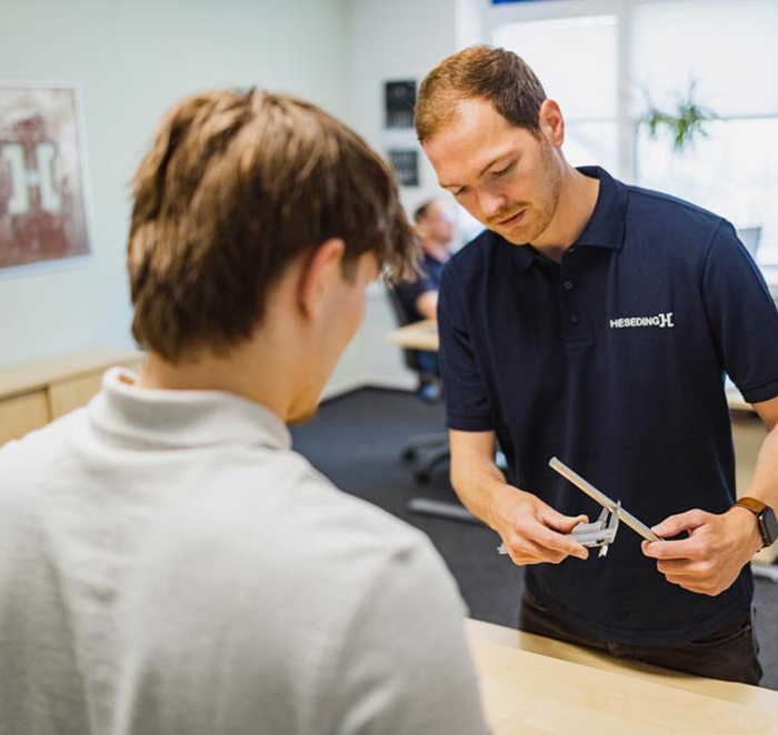 Auf dem Foto sieht man einen Heseding-Mitarbeiter, der einem Kunden im Showroom ein Werkzeug &ndash; vermutlich eine Schieblehre &ndash; erkl&auml;rt. Die beiden stehen an einem hellen Beratungstisch, im Hintergrund ist das moderne B&uuml;ro mit weiteren Mitarbeitenden zu sehen. Die Szene vermittelt kompetente, pers&ouml;nliche Beratung und zeigt, dass Heseding seinen Kunden fachkundigen Service auf Augenh&ouml;he bietet.